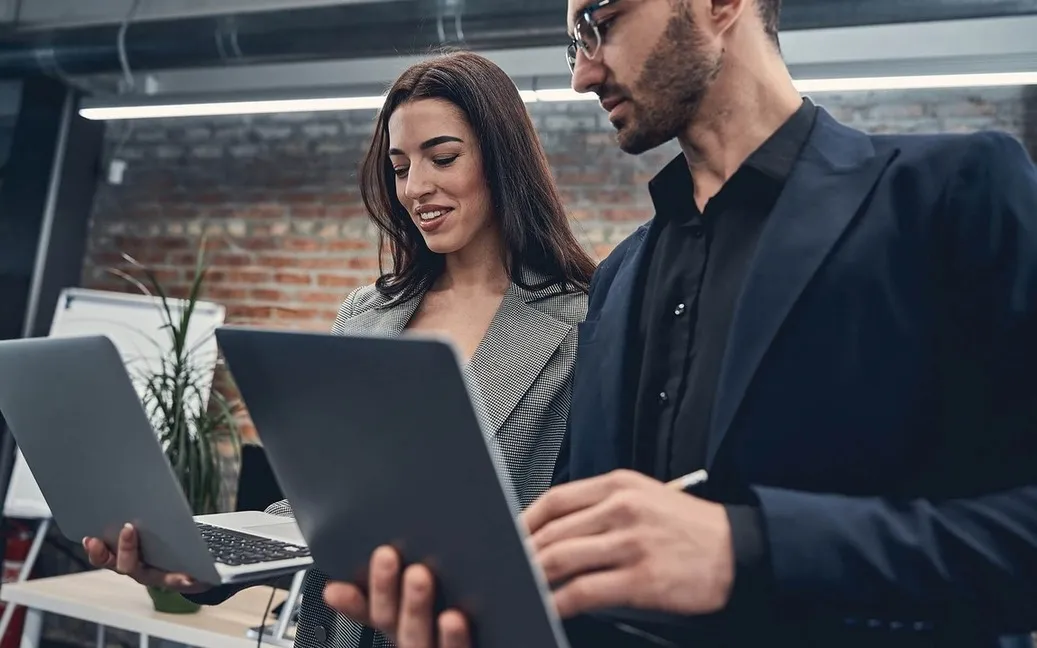 Dos profesionales de negocios de pie, ambos con laptops en mano, discutiendo algo con sonrisas en una oficina moderna.