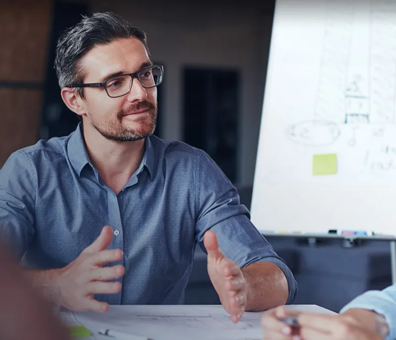 Un profesional con gafas sentado en una mesa, gesticulando mientras habla en una reunión, con una pizarra de fondo.